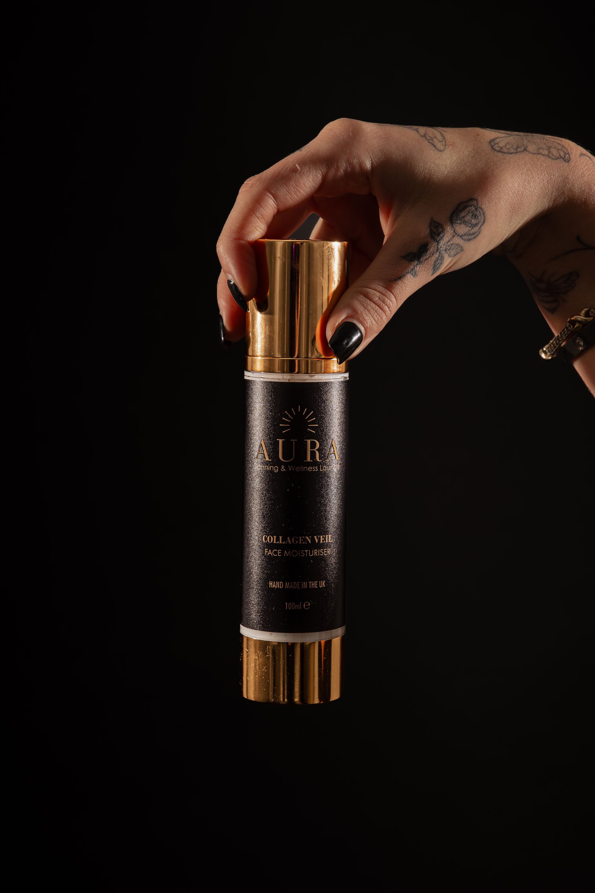 Tattooed hand holding a black-and-gold cylindrical bottle against a dark background