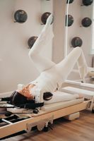 Woman doing Pilates on a reformer, legs in straps, arms raised. Neutral setting, beige tones.