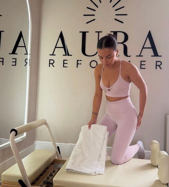Woman doing Pilates on a reformer, legs in straps, arms raised. Neutral setting, beige tones.