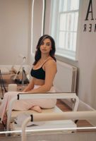 Woman doing Pilates on a reformer, legs in straps, arms raised. Neutral setting, beige tones.
