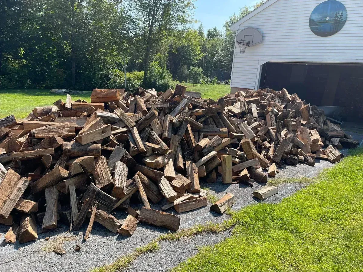 Large pile of chopped firewood stacked beside a white house and garage in a sunny yard.