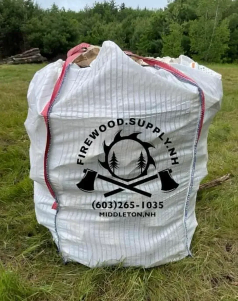 White firewood bag with red straps and black logo, standing outdoors in a grassy field.