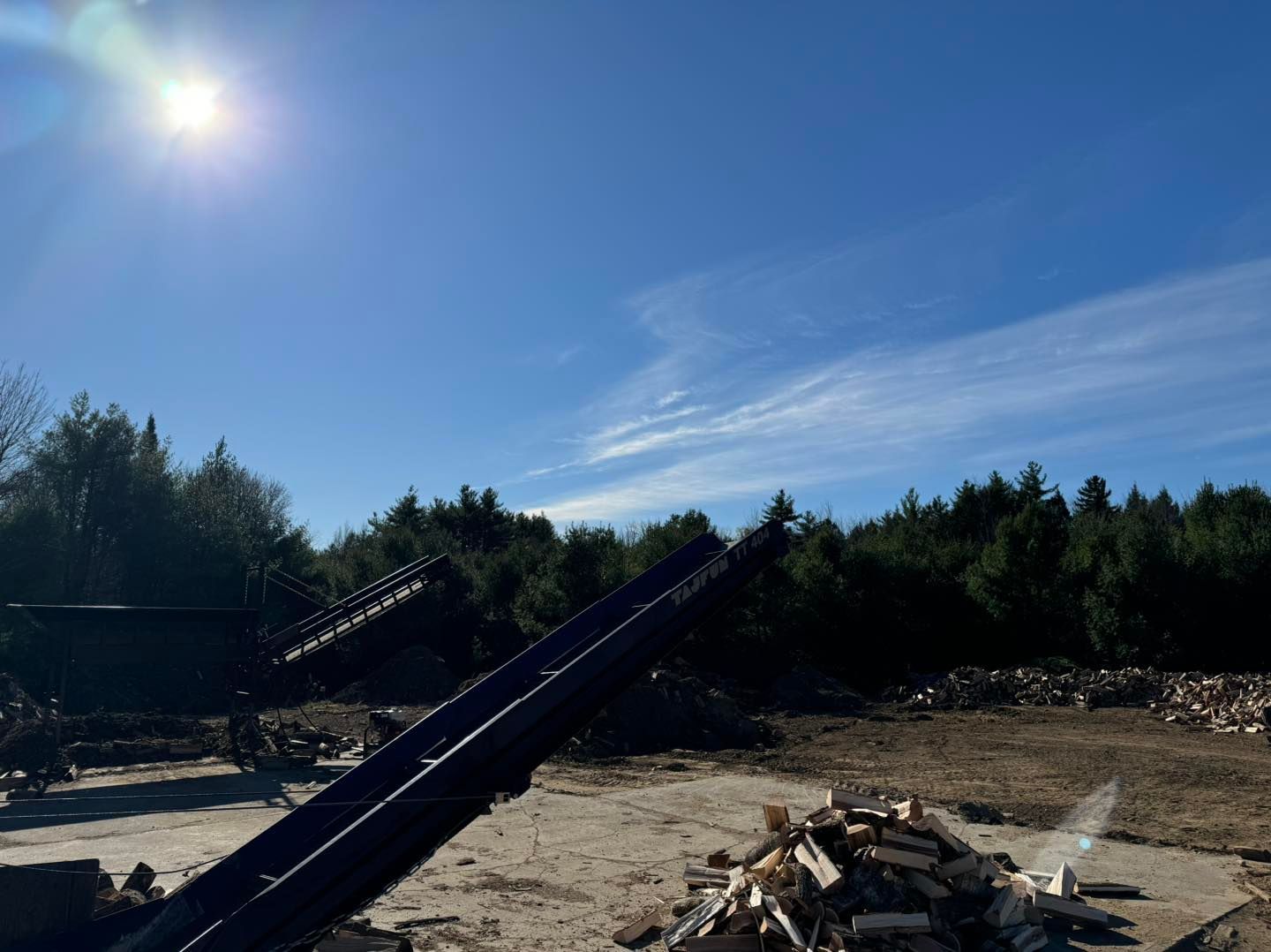 Sunny industrial yard with two conveyor belts and a pile of wood debris under a blue sky