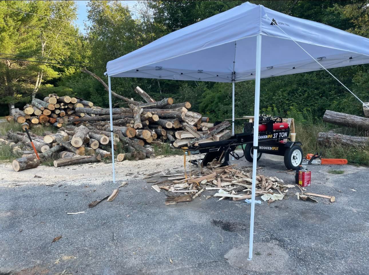 White pop-up canopy beside stacked firewood and a red trailer in a gravel yard