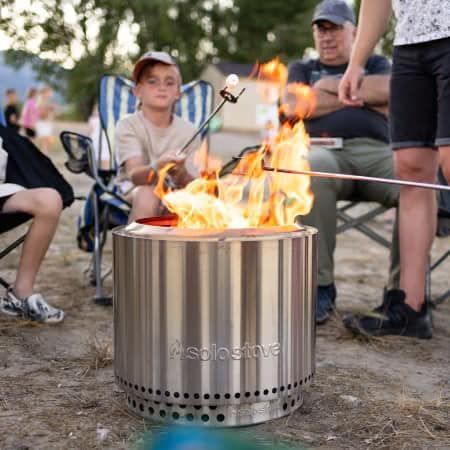 People gathered around a stainless steel fire pit with flames outdoors at dusk