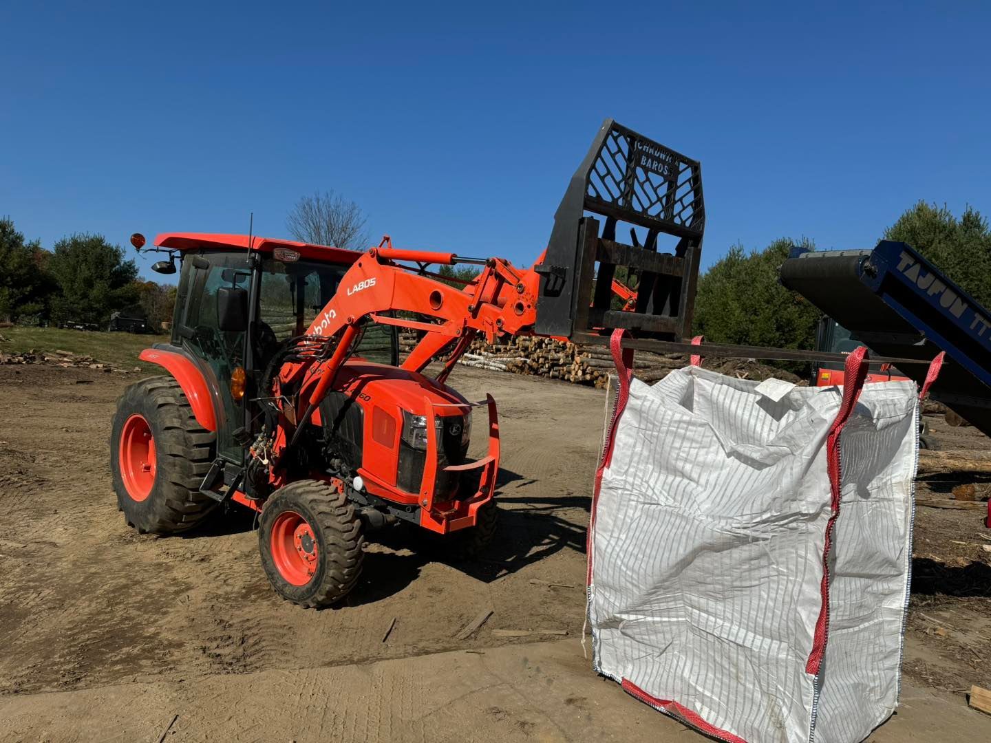 Orange tractor with front loader lifting a white bulk bag in a dirt lot under a clear blue sky