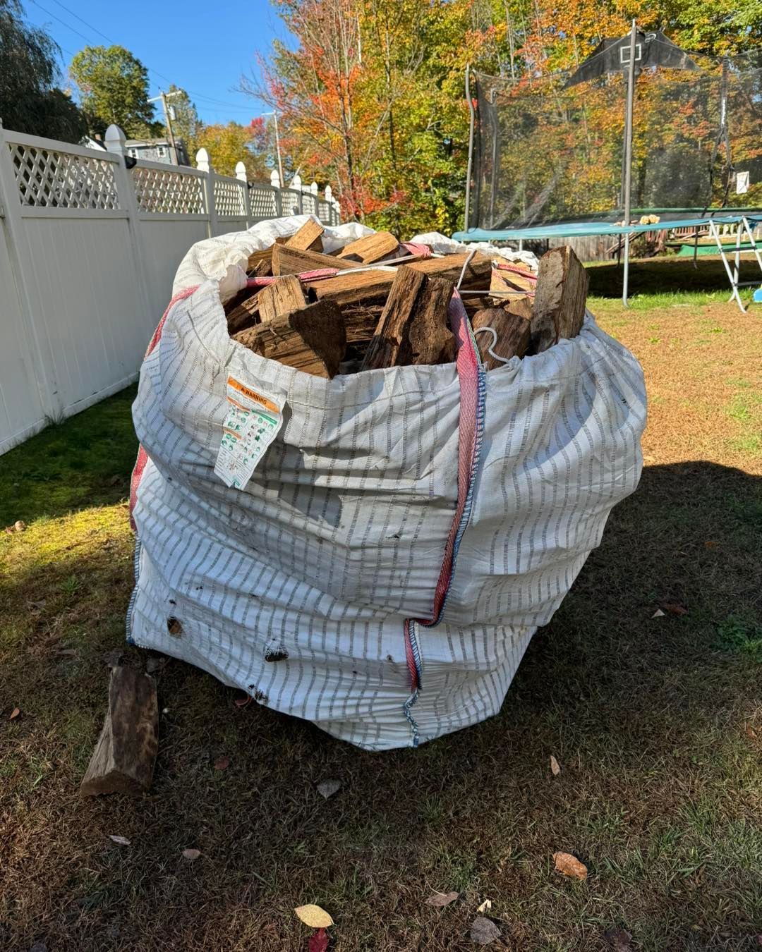 Large white bulk bag filled with cut wood pieces in a backyard, beside a white fence and autumn trees