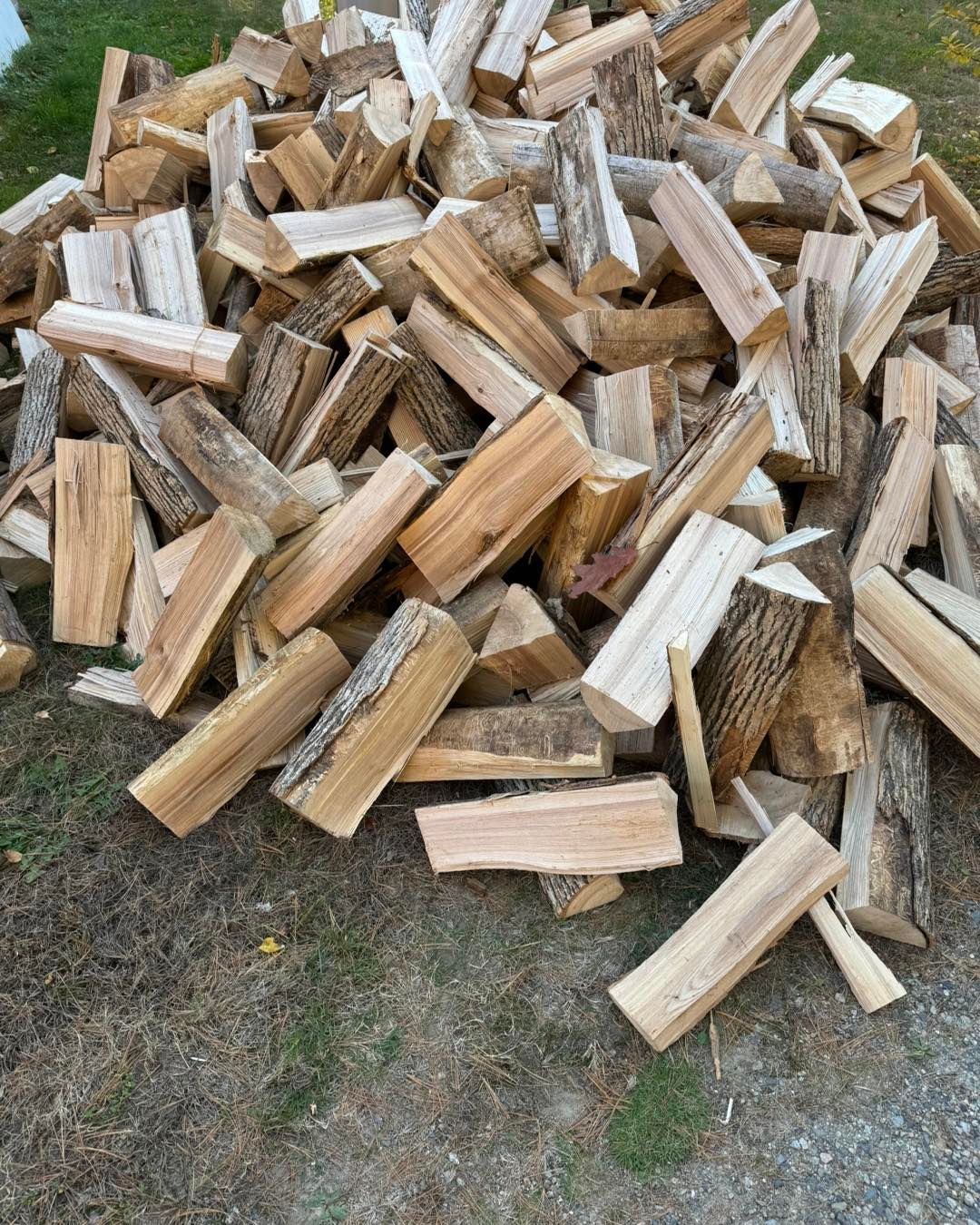 Pile of chopped firewood on gravel, with light brown split logs and a few green weeds nearby