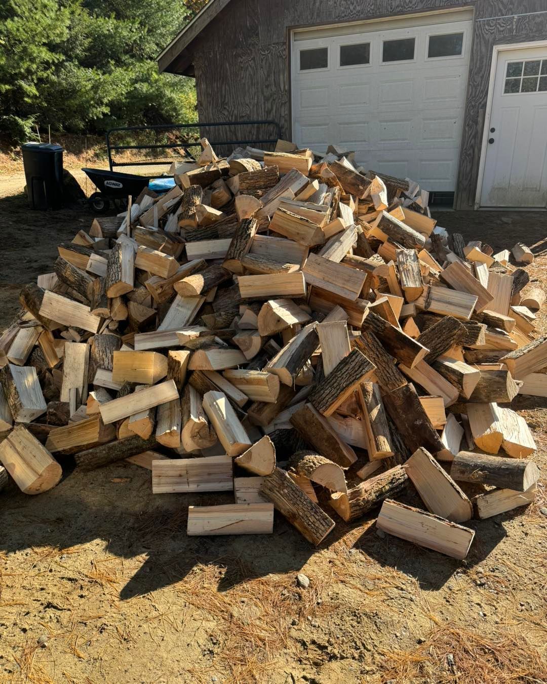 A large pile of split firewood stacked on dirt in front of a garage and house.