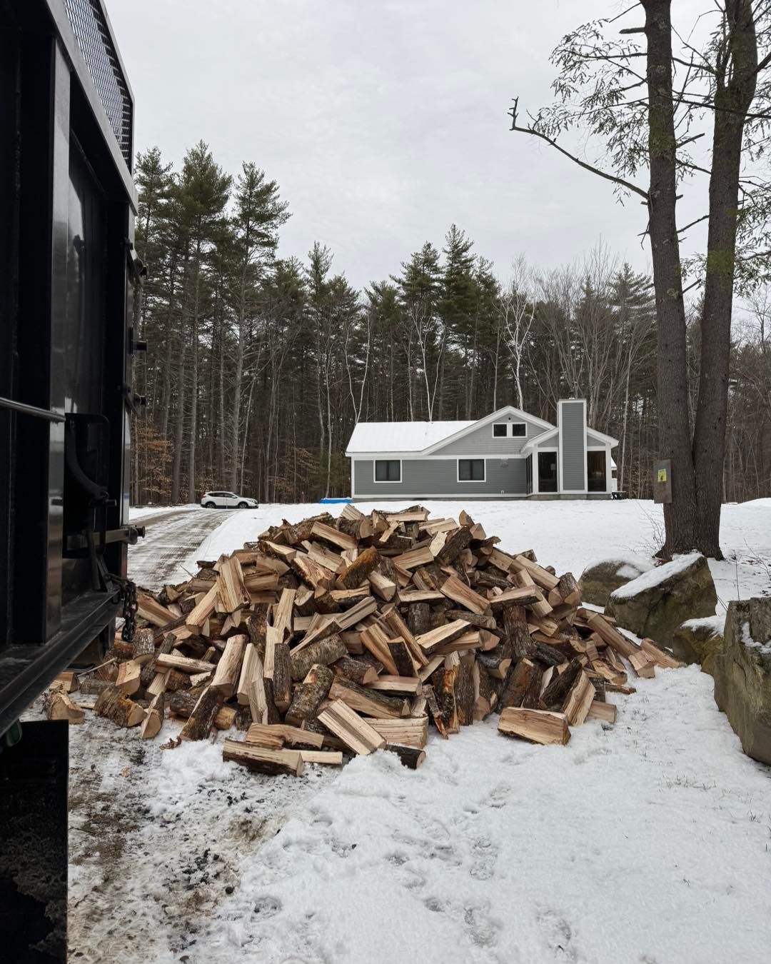 Snowy yard with a large woodpile near a house and forest in the background