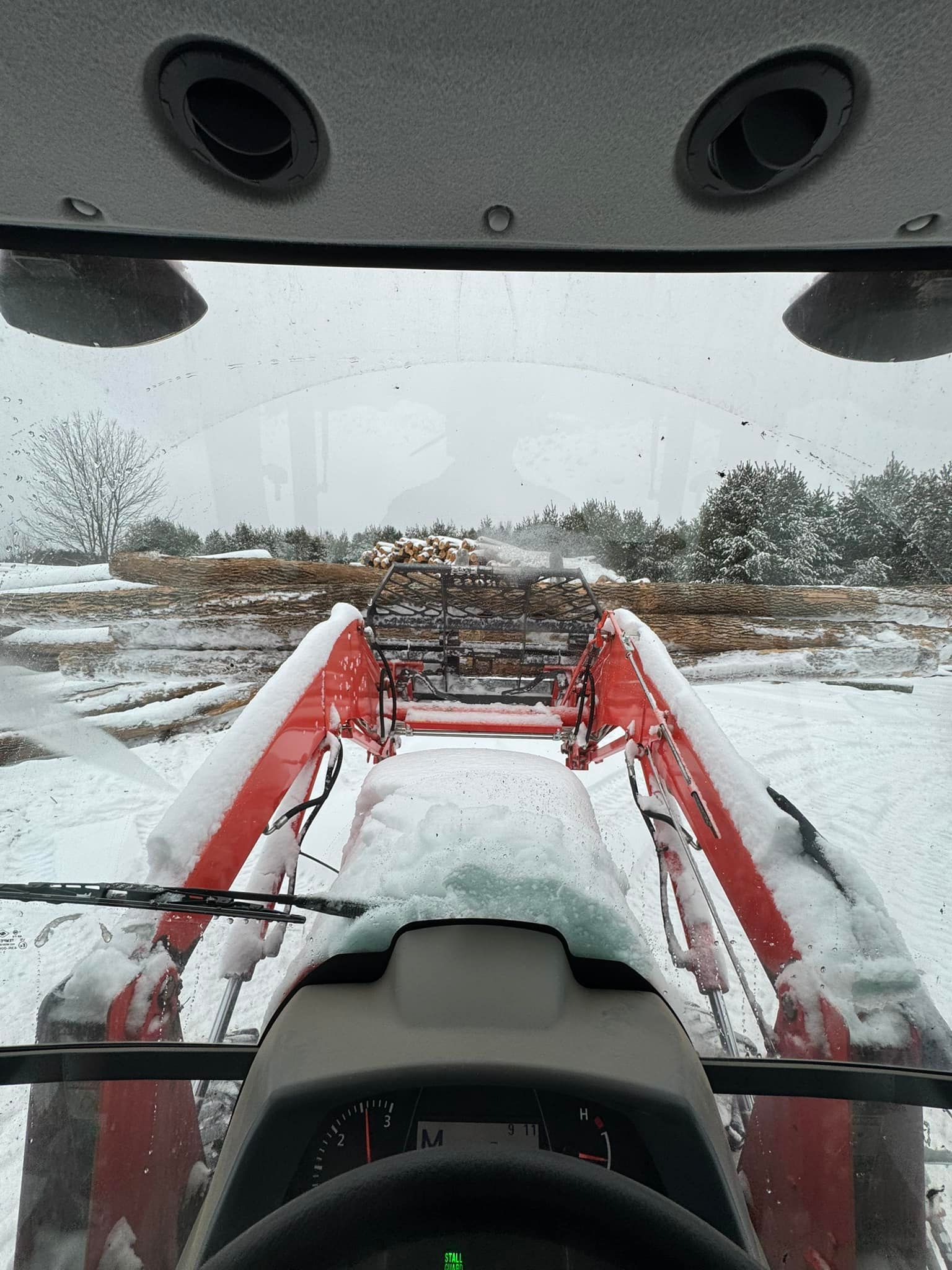 View from a tractor cab over red loader arms in a snowy field, facing a brush pile.