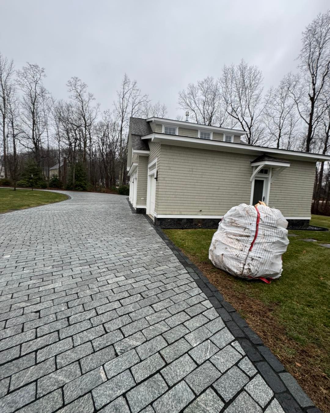 Stone driveway beside a beige house, with a large white yard waste bag on the lawn.