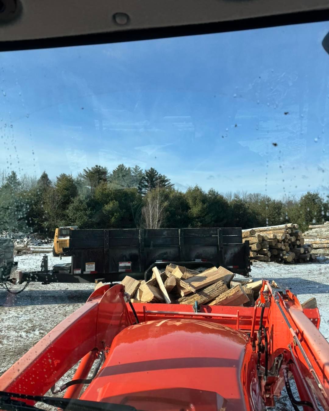 Red tractor cab facing a wood pile near black dump trailers on a gravel lot under a blue sky