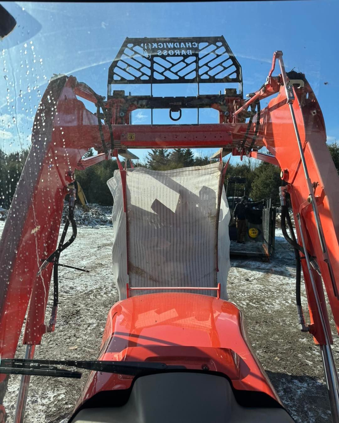 View from inside a red tractor with front loader and protective cab screen, parked on gravel under a blue sky