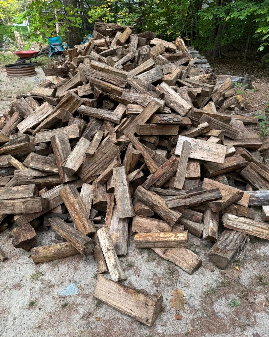 Pile of split firewood logs stacked outdoors on a gravel surface beside green trees