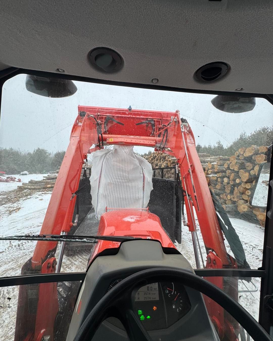 View from a red tractor driving through snow beside stacked logs in a forest clearing