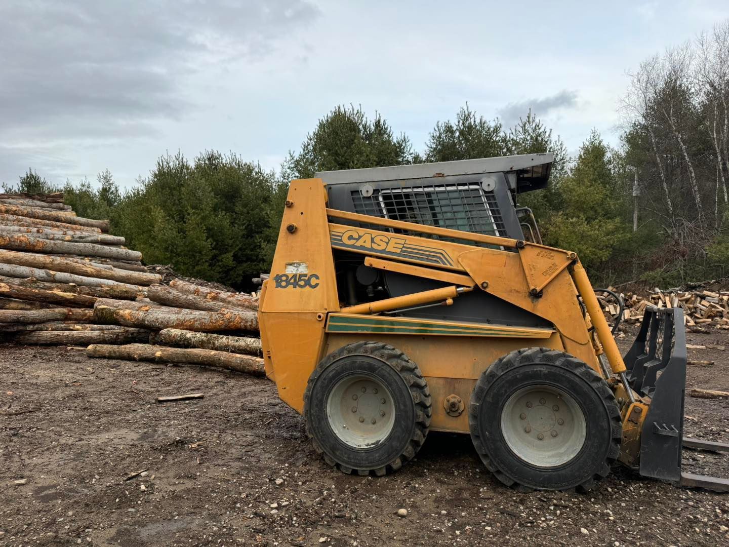Yellow skid steer loader beside stacked logs in a muddy forest clearing