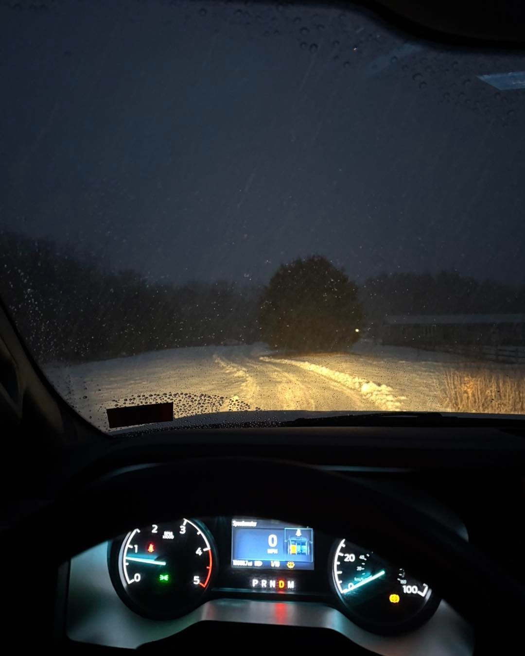 Driver’s view of a snowy road at night through a rain-speckled windshield, dashboard lights glowing