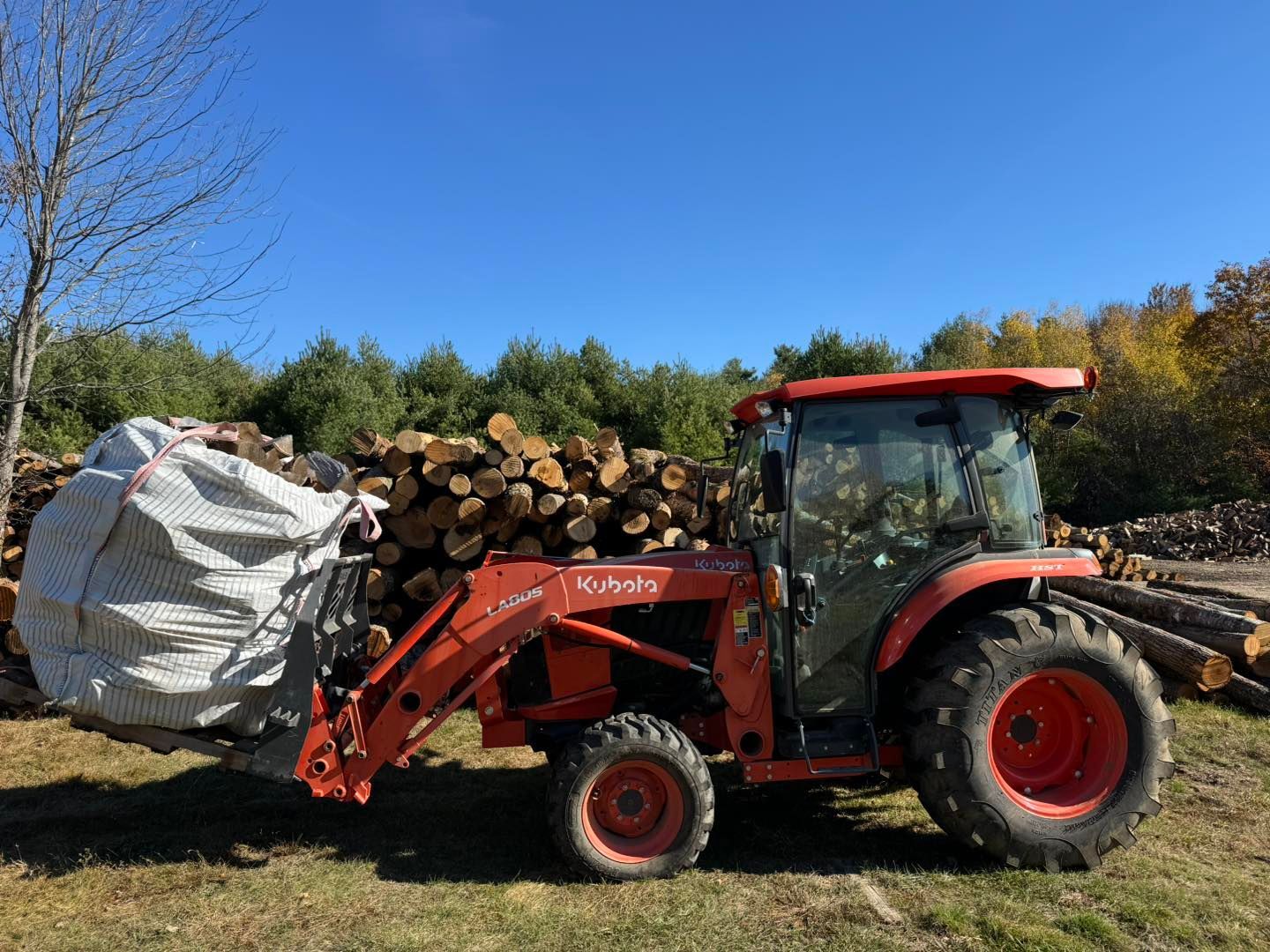 Red tractor carrying a large bag and stacked logs in a field under a blue sky