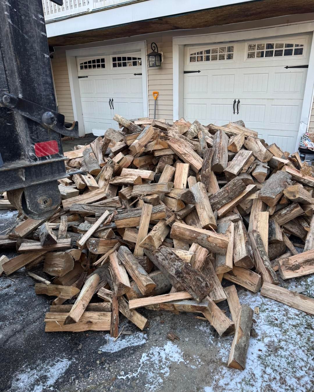 Large pile of chopped firewood stacked on a driveway in front of two white garage doors.