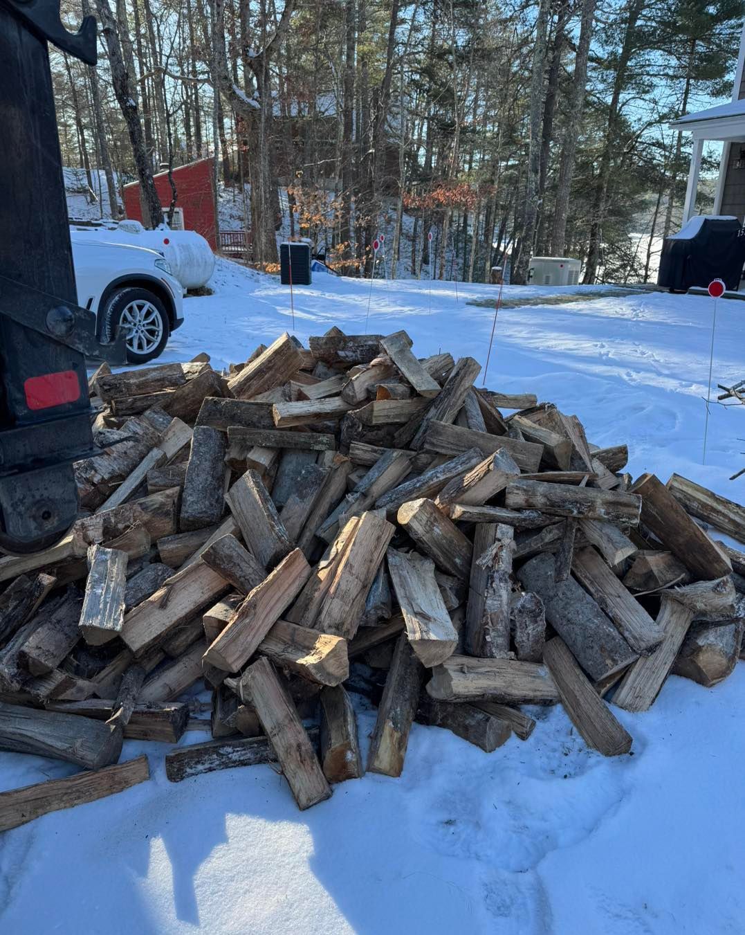 Pile of split firewood on snow in a wooded driveway, with parked vehicles in the background.