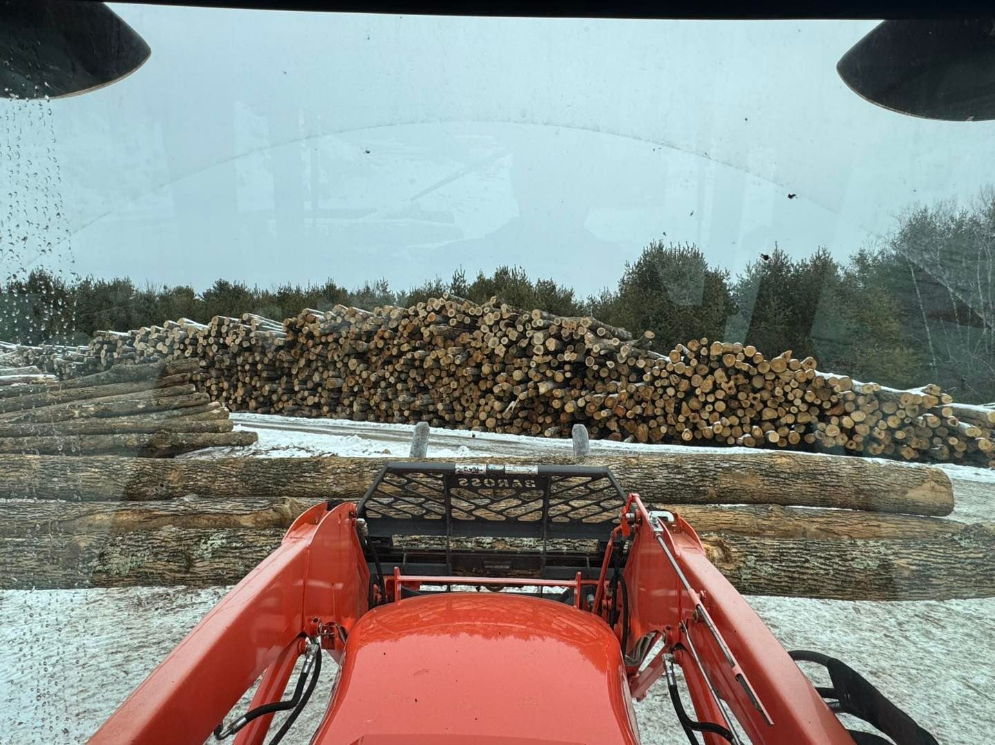 View from a red tractor cab toward stacked logs in a snowy yard