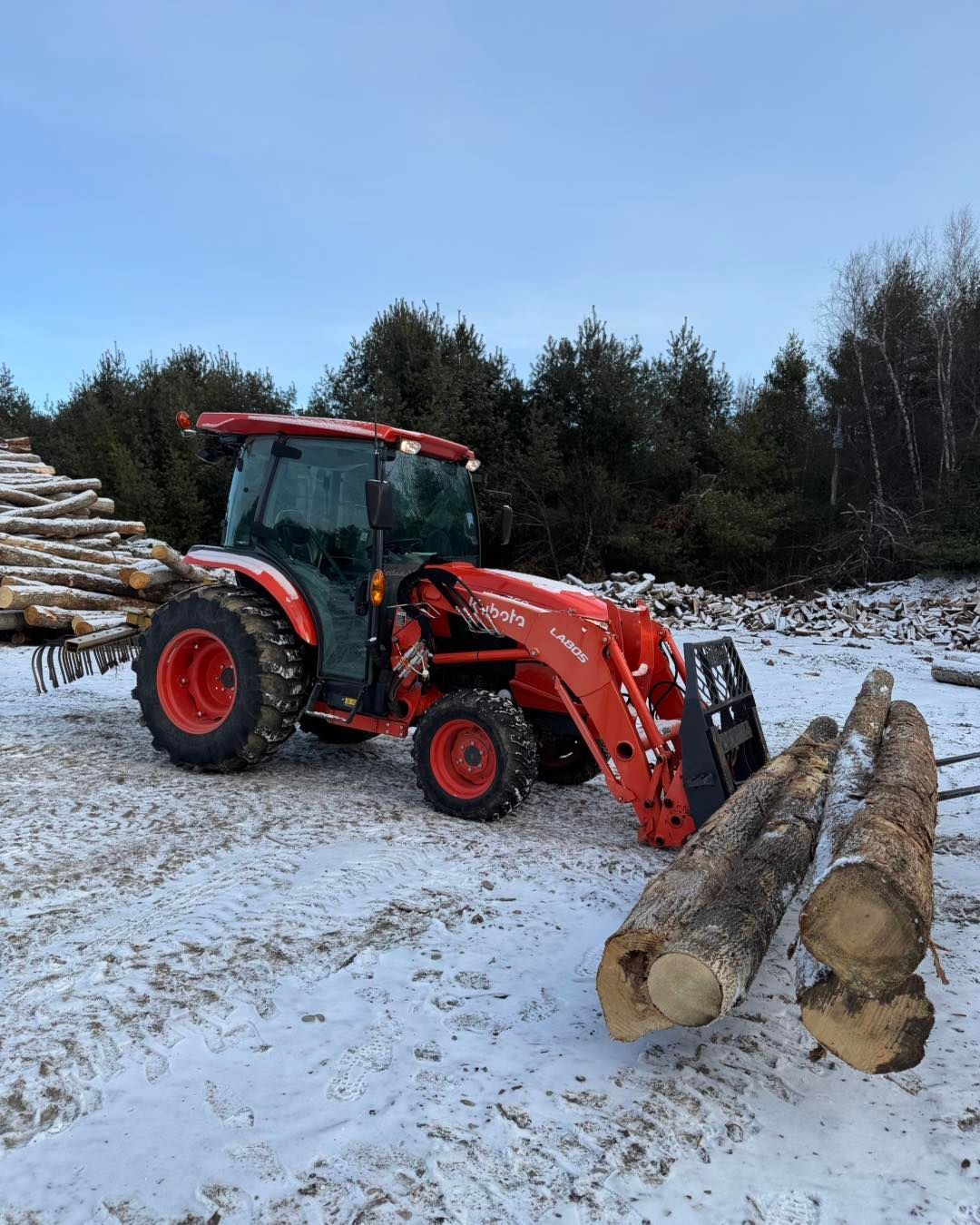 Orange tractor with front loader moving logs in a snowy forest clearing