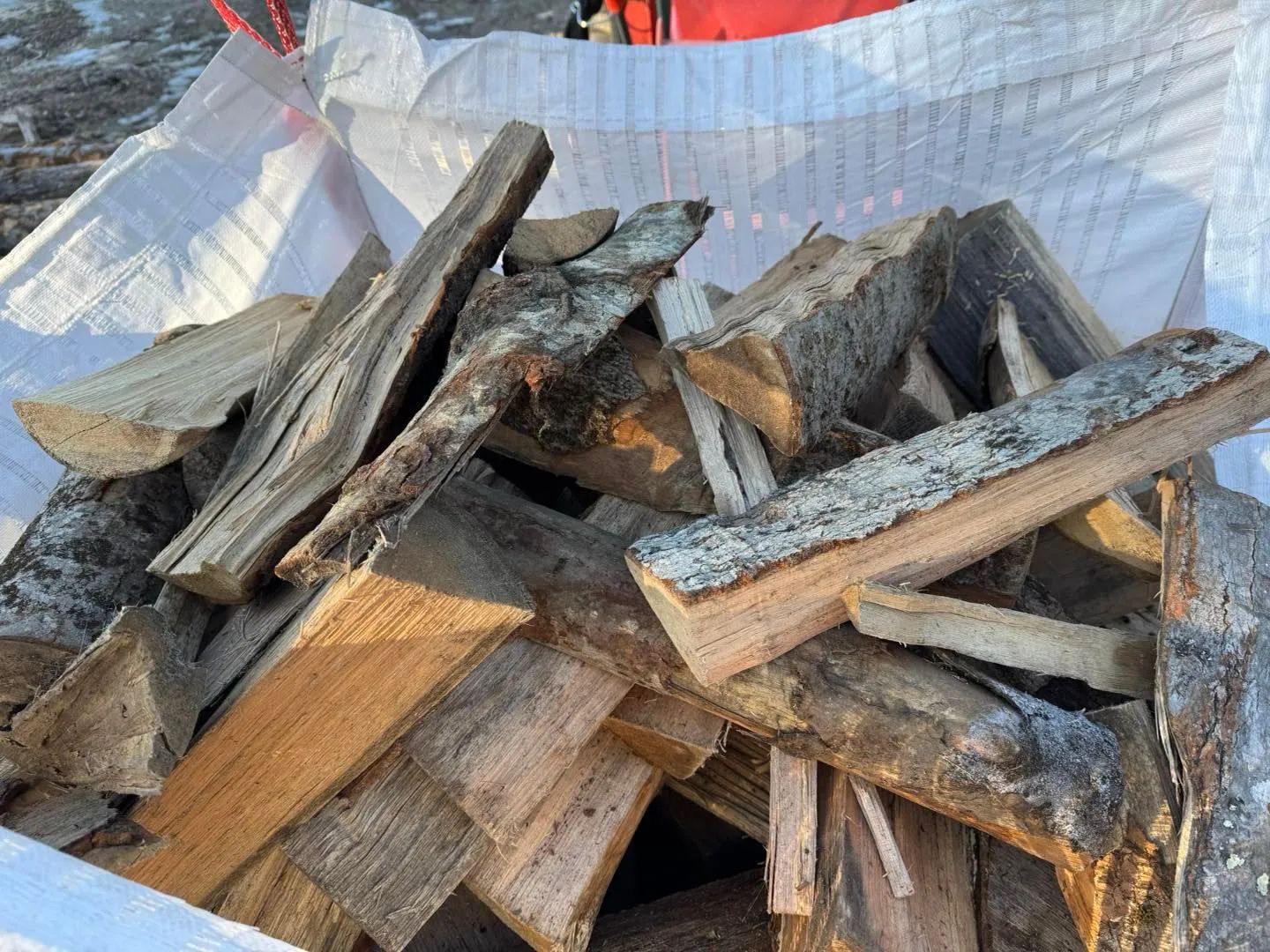 Firewood in a white bag, showing cut wood with various bark colors, and textures.