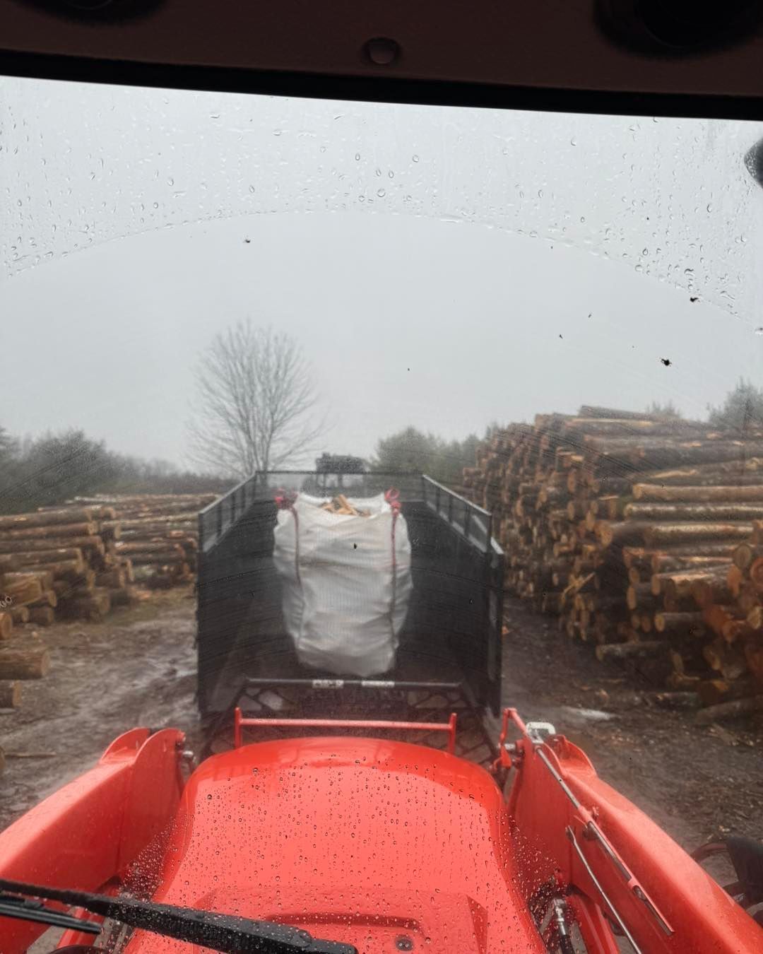 View from a red tractor driving between stacked logs on a rainy forest road