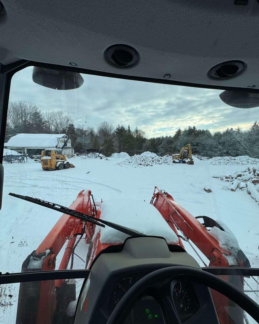 View from a tractor cab over red loader arms in a snowy yard with a barn and trees in the distance