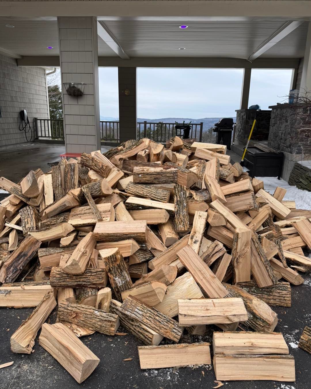 Large pile of chopped firewood stacked under a covered patio with an outdoor view beyond.