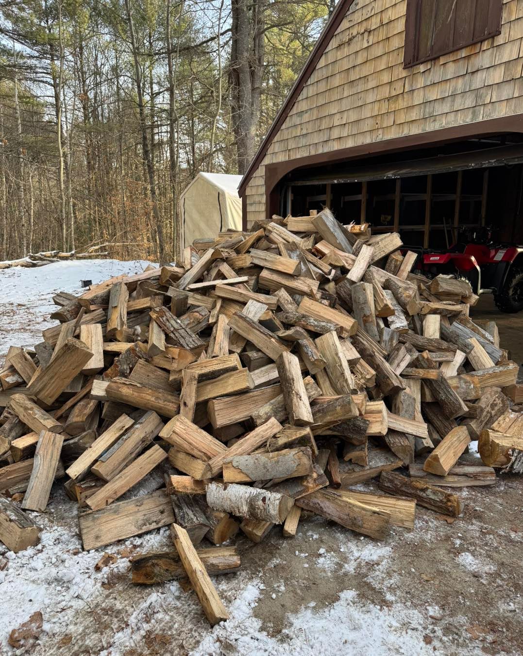 Large pile of chopped firewood stacked beside a garage in a snowy yard.