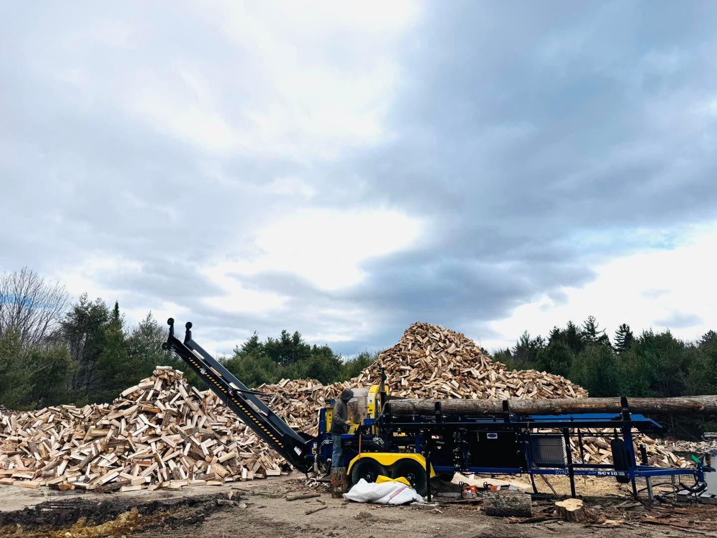 Wood chipper unloading logs into a large pile at an outdoor recycling yard under cloudy sky