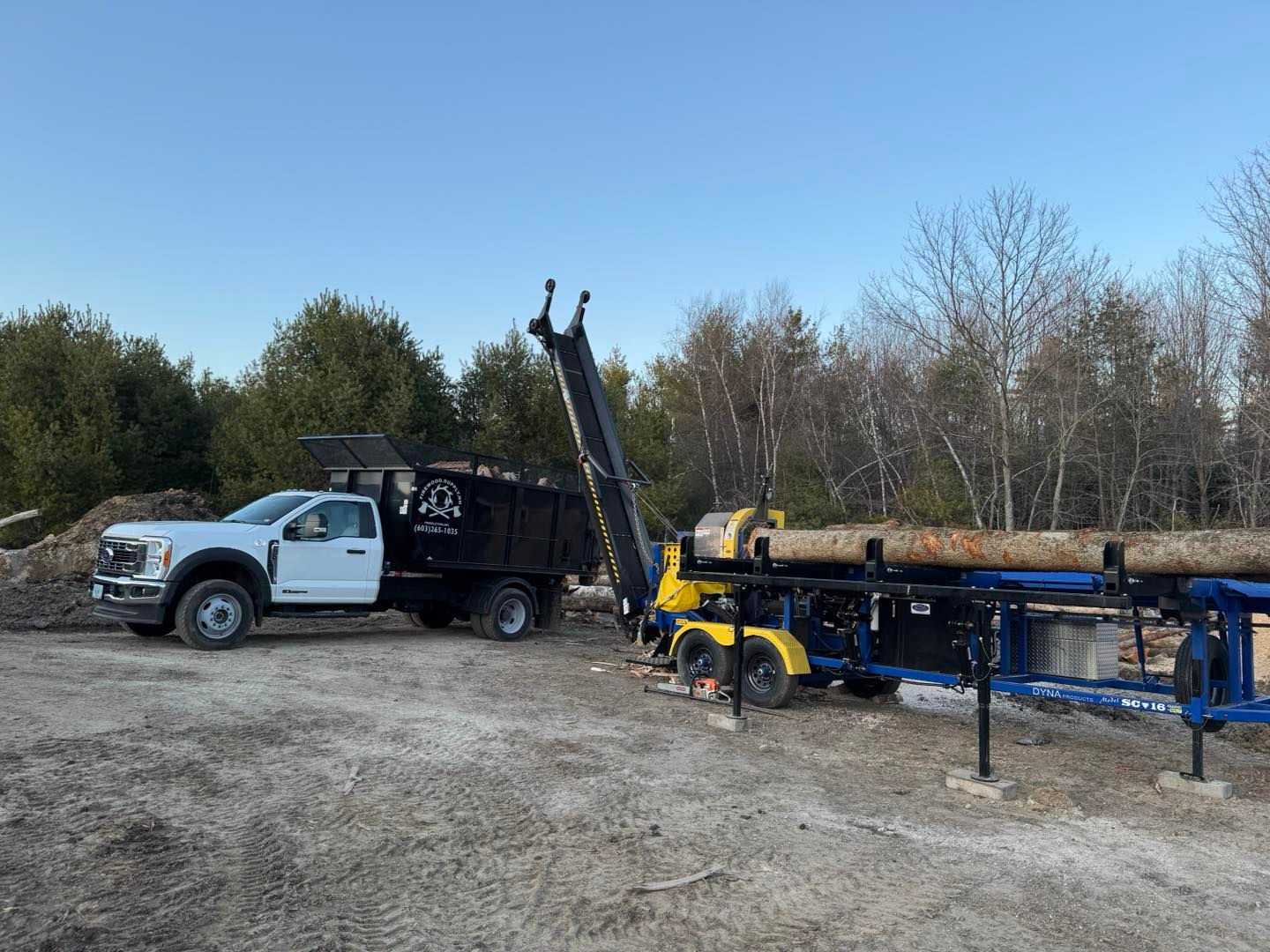 White work truck and black dump trailer beside yellow farm equipment on a gravel lot under a clear sky