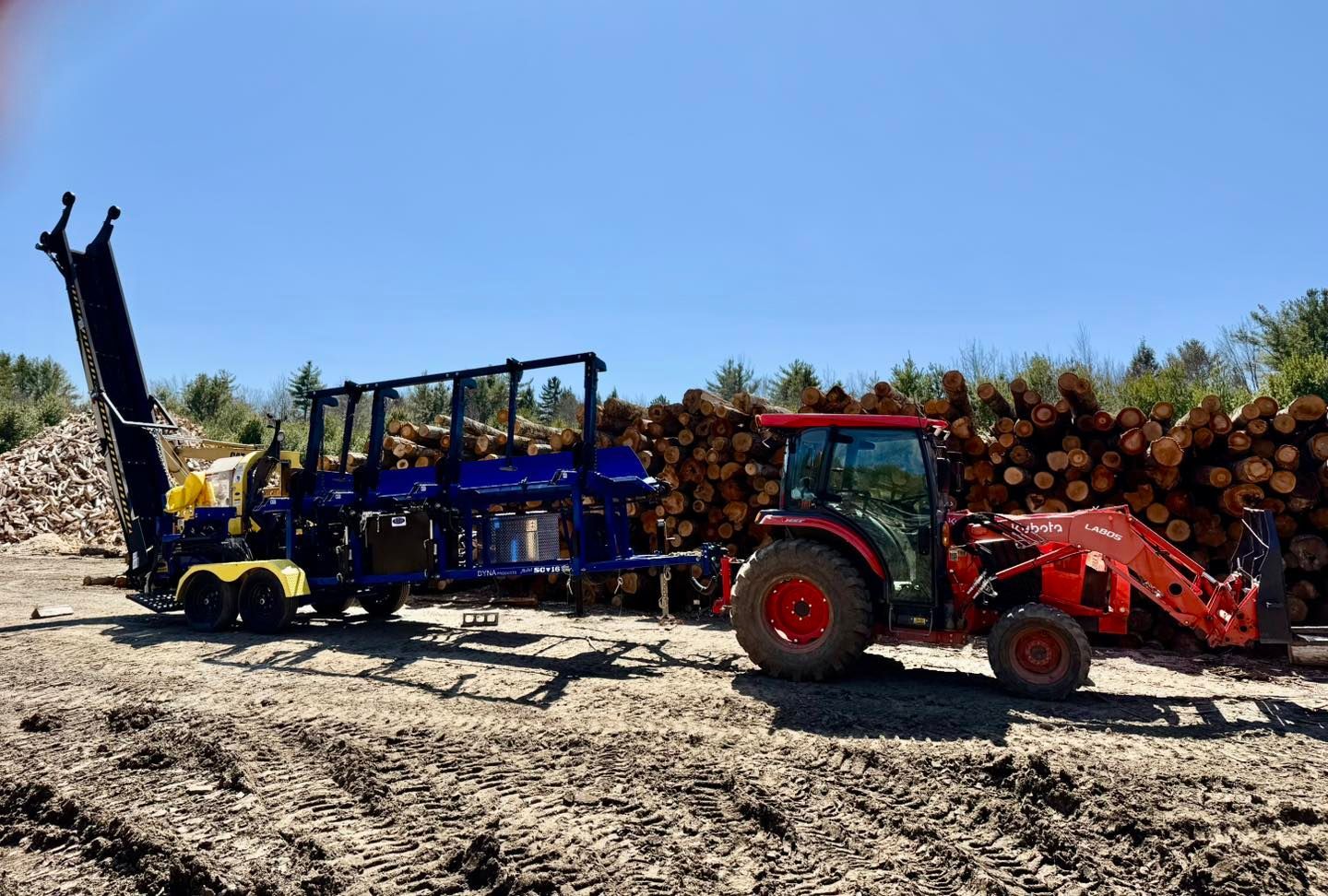 Red tractor with front loader beside blue logging trailer on rocky ground under a clear sky