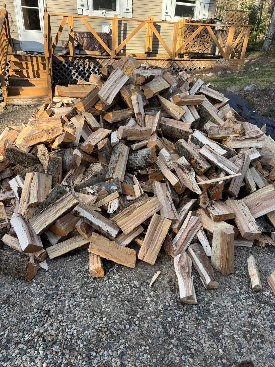 Pile of split firewood stacked on gravel in a yard, with a wooden porch railing in the background