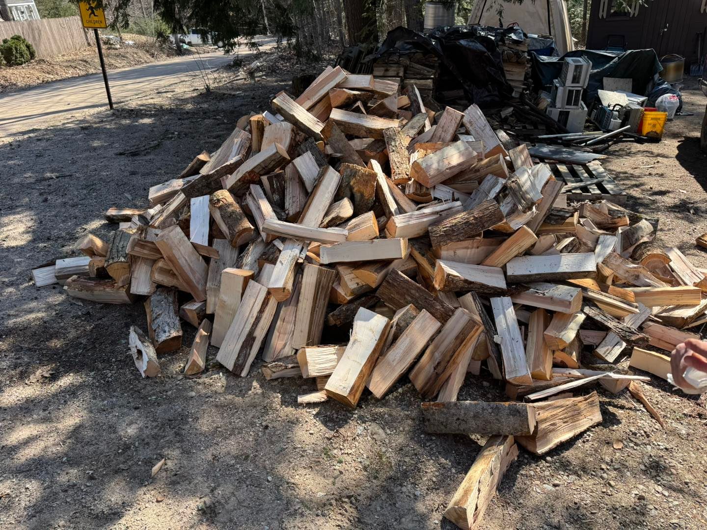 Pile of chopped firewood stacked outdoors on dry ground, with tools and yard equipment in the background.