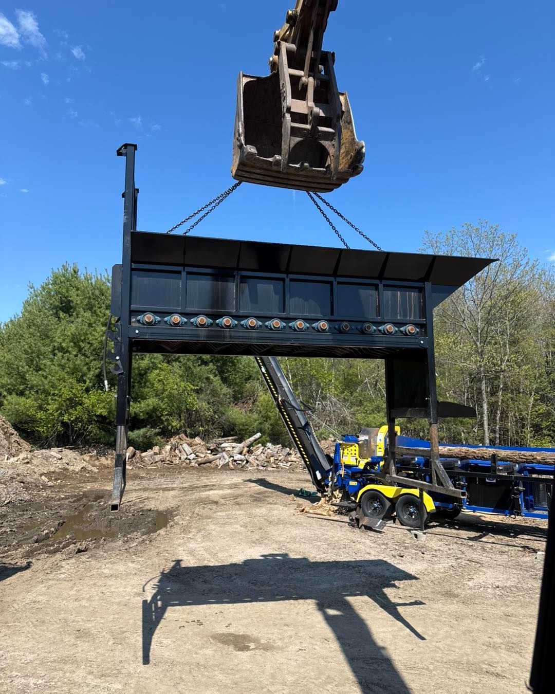 Excavator lifts a large black metal hopper beside a yellow utility truck on a dirt site under a blue sky