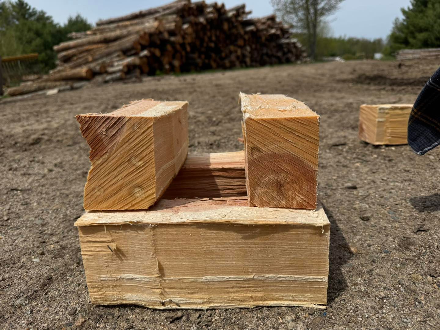 Stacked split firewood on dirt, with a pile of logs in the background.