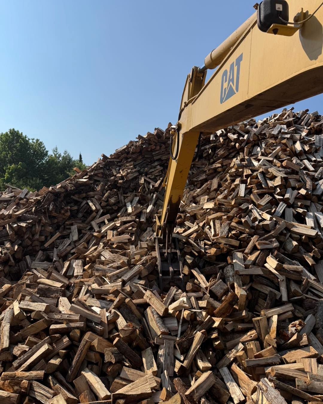 Excavator arm digging into a large pile of wood debris under a clear blue sky