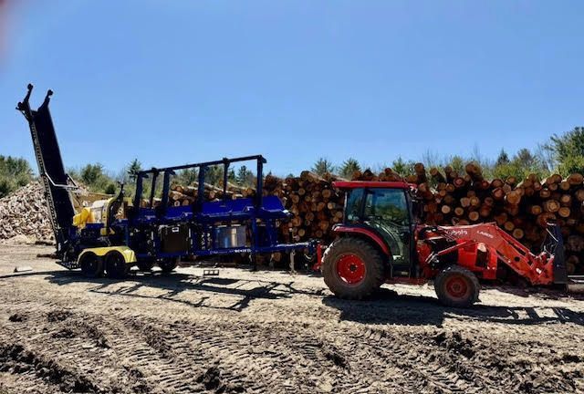Two tractors with logging equipment on a gravel lot, stacked logs and blue sky behind them