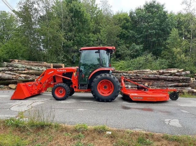 Red tractor with loader and mower parked beside stacked logs in a wooded roadside area.