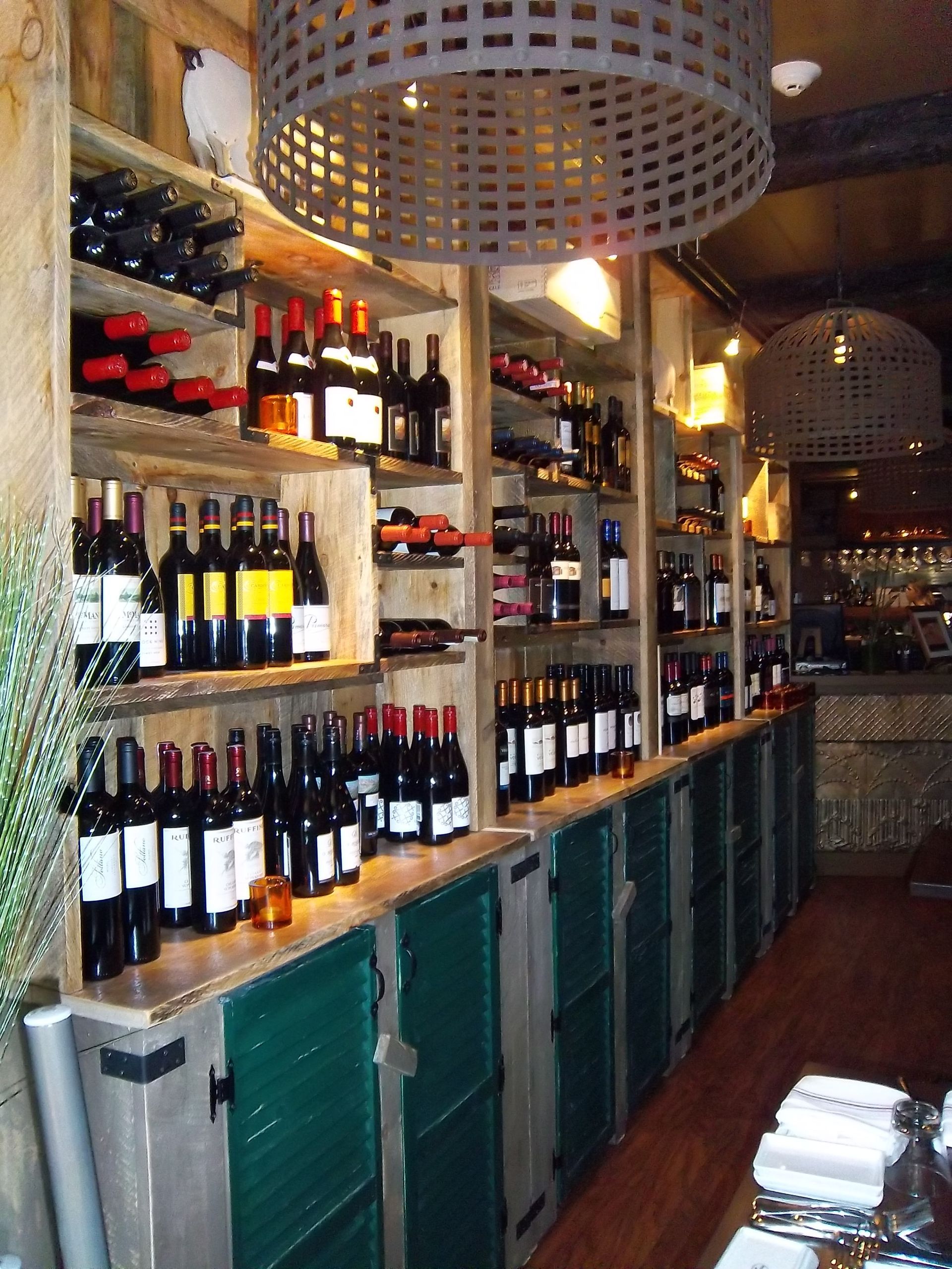 A rustic wine shop featuring wooden shelves packed with wine bottles above green storage cabinets and wicker lamps.