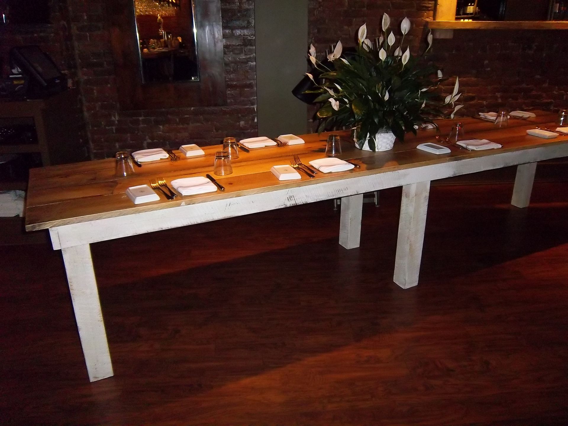A long wooden dining table with a white base, set for guests, with a large white flower centerpiece in a dimly lit room.
