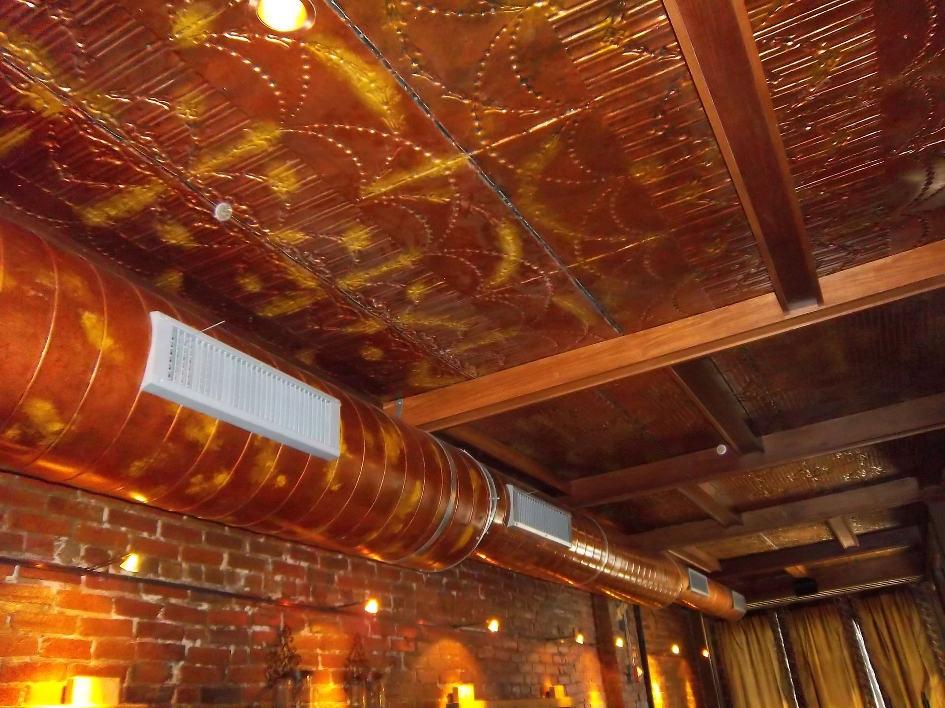Copper-toned ceiling with patterned tiles and exposed beams above a cylindrical ventilation duct and brick wall.