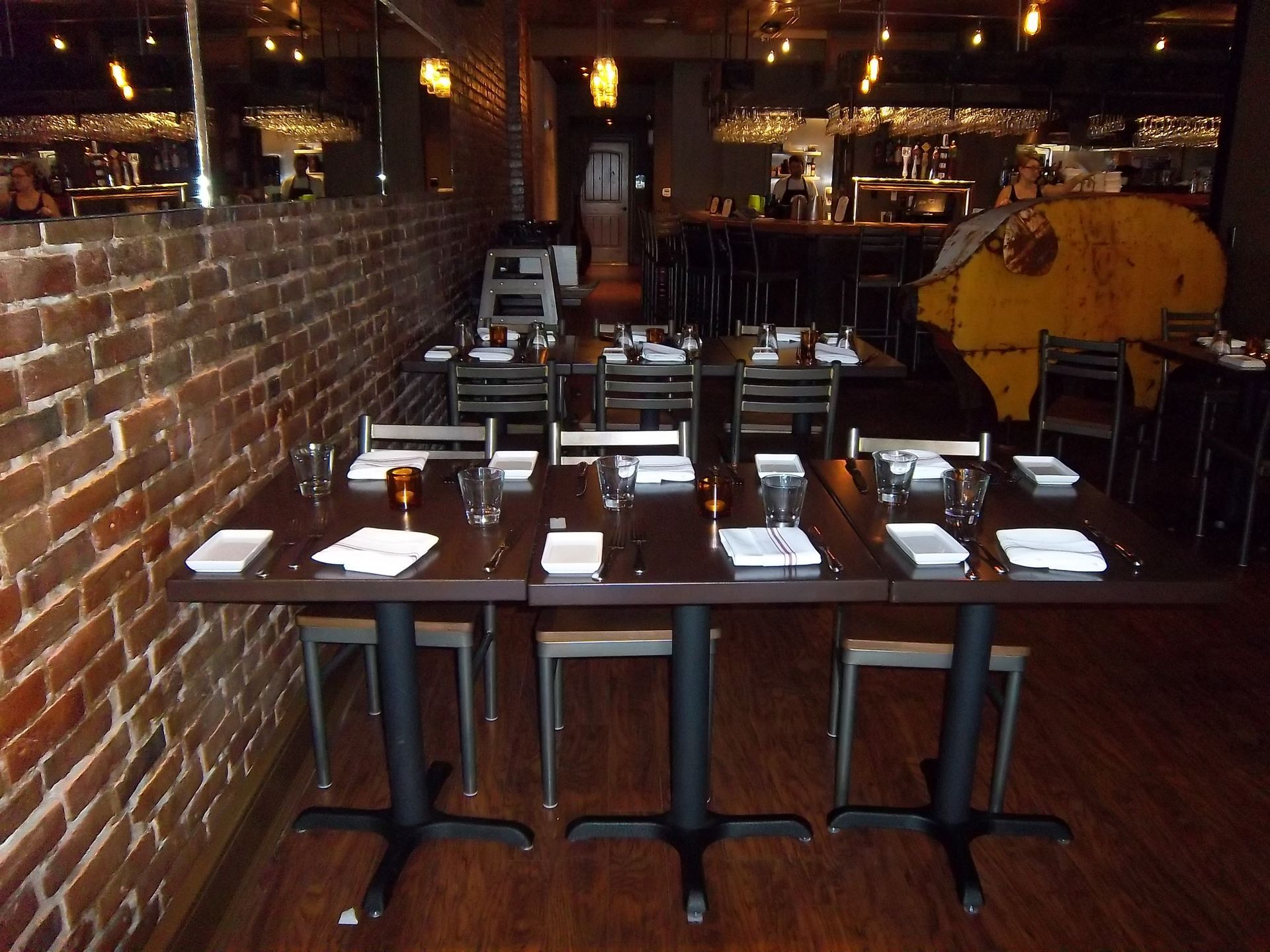 A series of dark wooden tables set for dining along an exposed brick wall inside a dimly lit restaurant.
