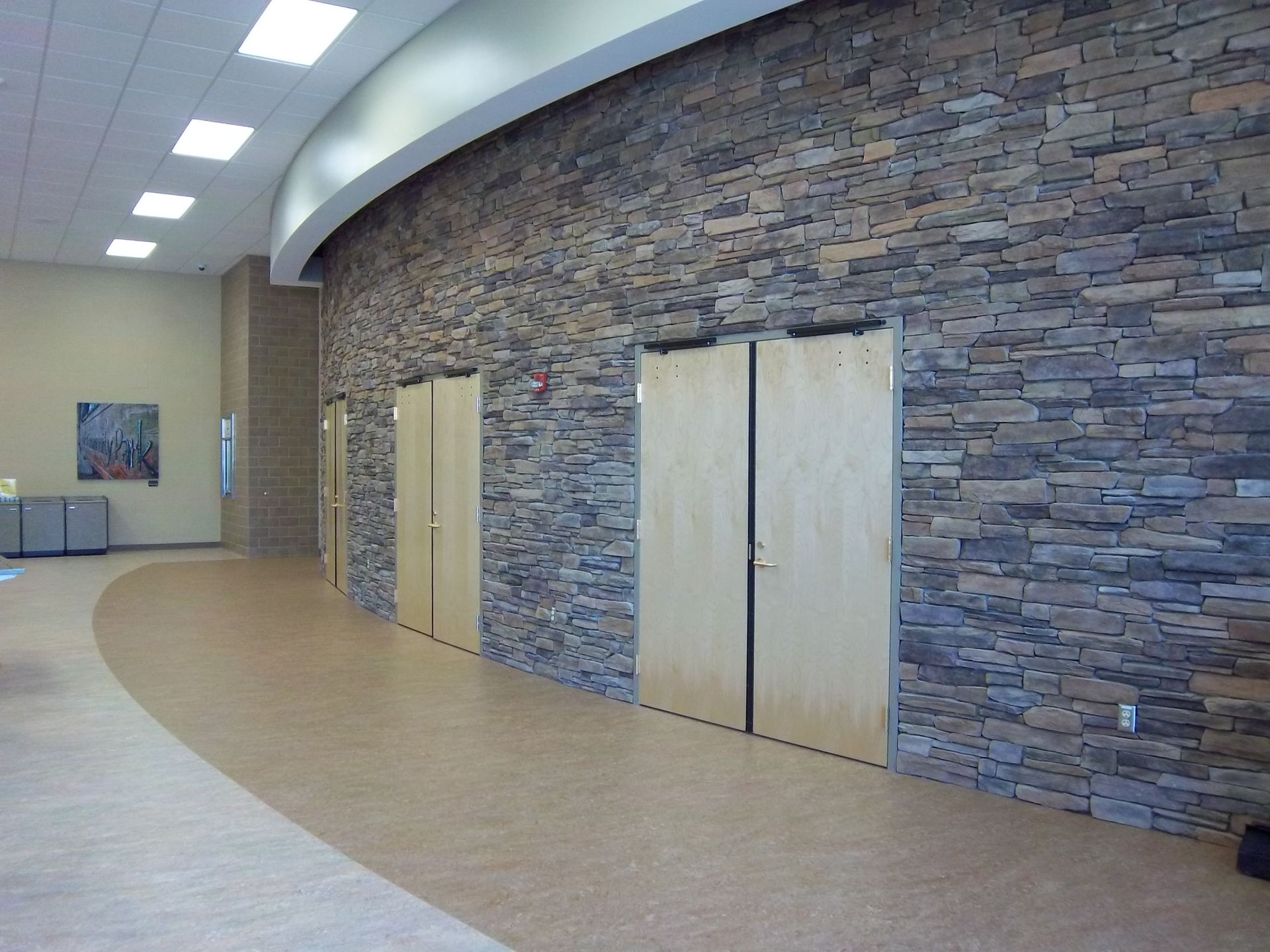 A hallway with a curved stone wall and light wood double doors under recessed ceiling lights.