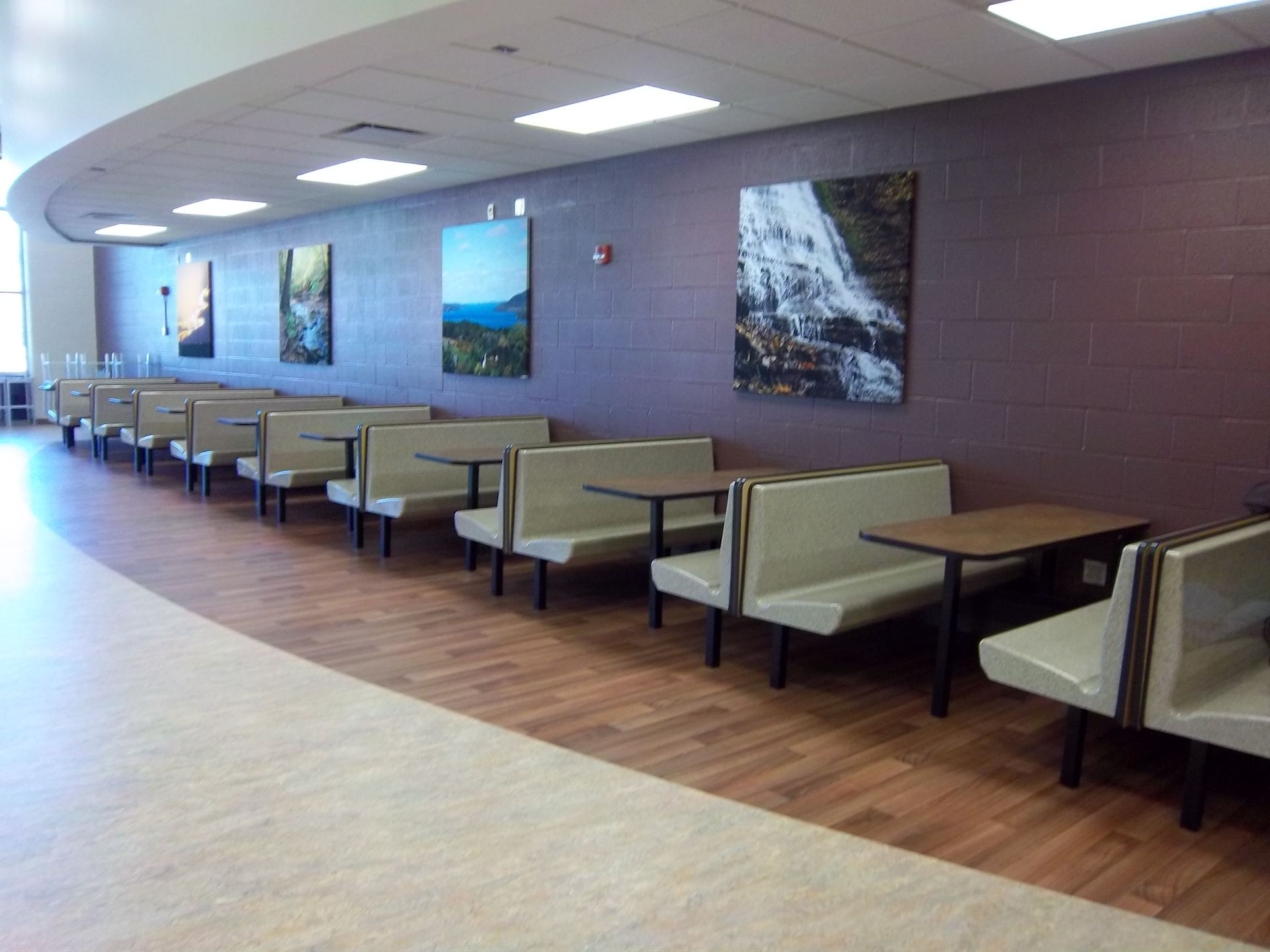 Empty cafe booths with beige seats and brown tables line a curved, purple-walled hallway with nature art prints.