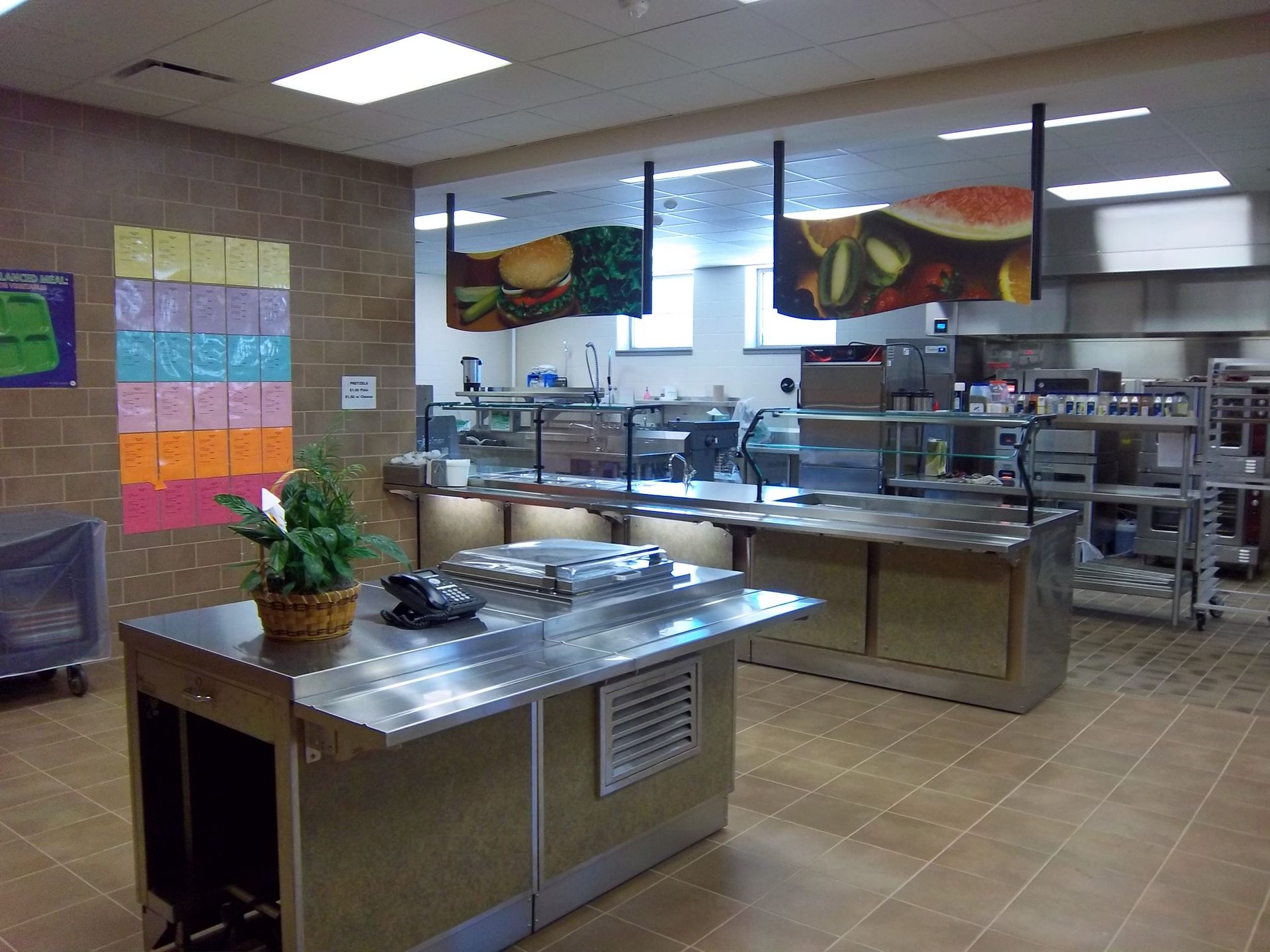 A school cafeteria serving line with a stainless steel counter, food stations, and colorful wall decorations.