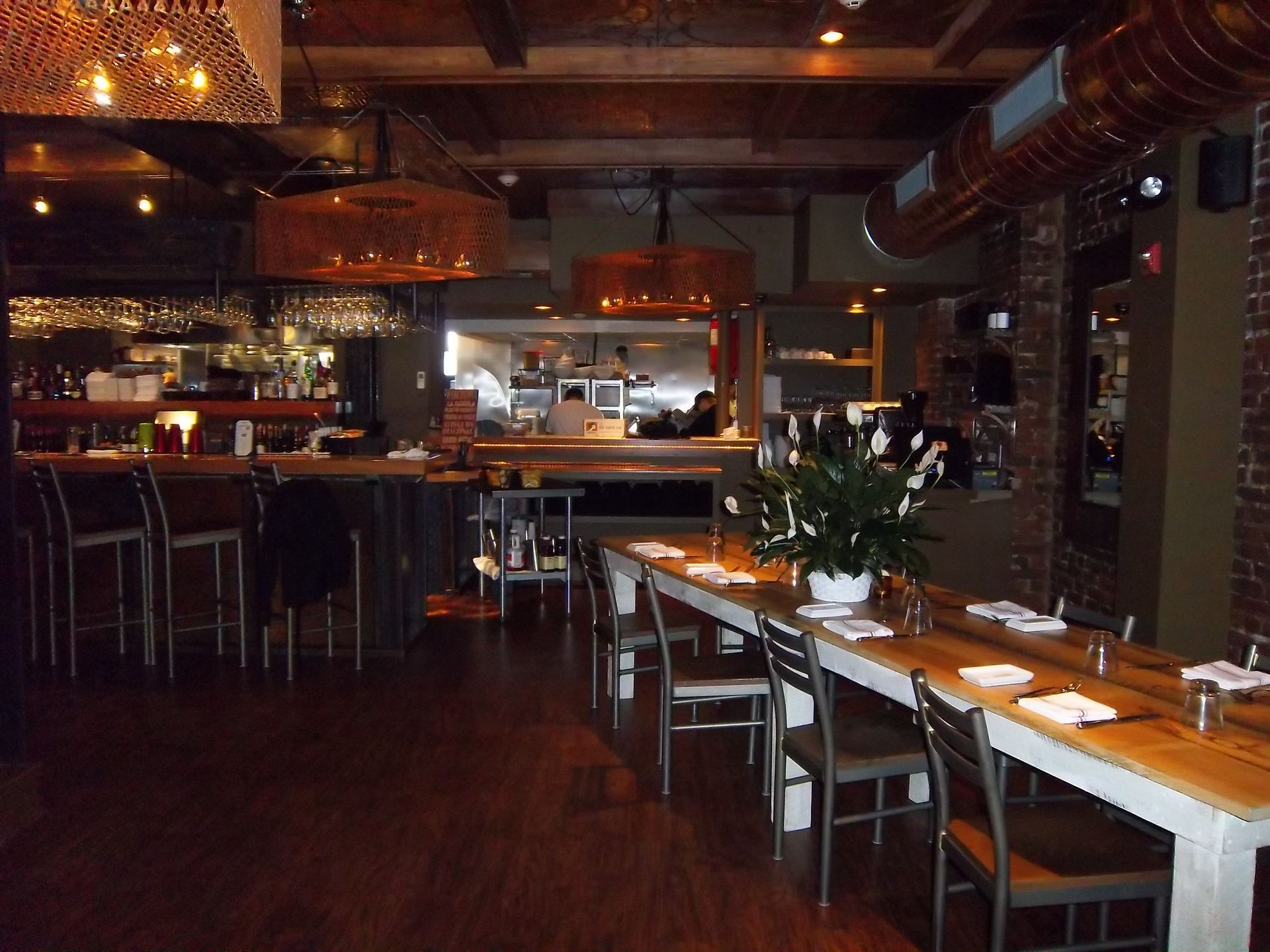A dimly lit restaurant interior with a long communal wooden table, chairs, a bar area, and warm hanging pendant lights.